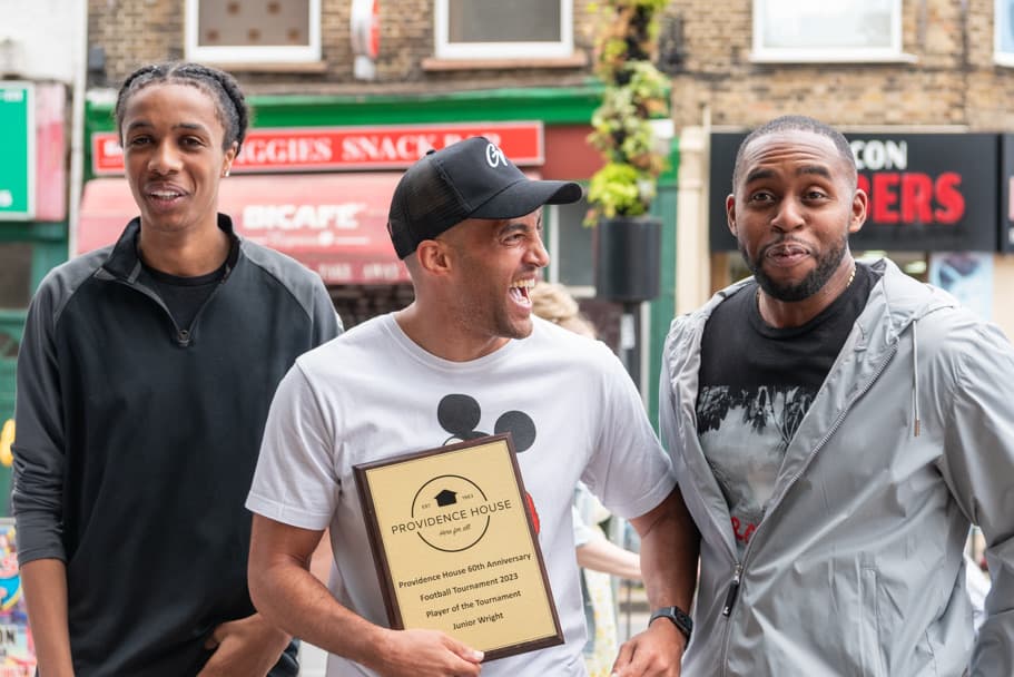 Three young men laugh together on a Battersea high street, one holding a plaque that reads Providence House 60th Anniversary Football Tournament 2023, Player of the Tournament