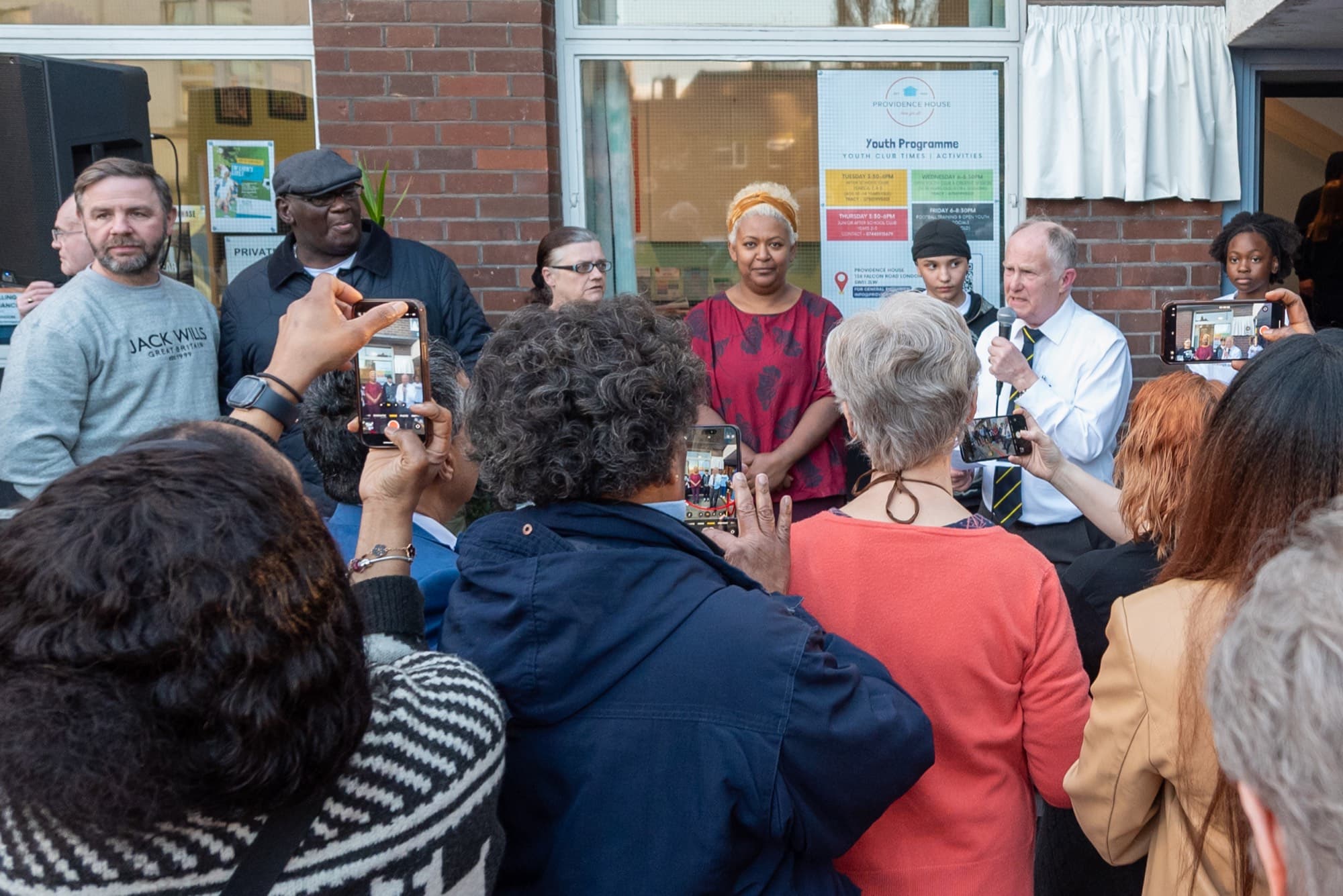 Neighbours, young people and supporters gather outside the Providence House entrance on Falcon Road as a speaker addresses them, many filming on their phones, with the youth programme poster in the window